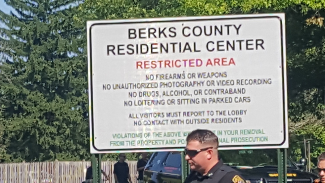 A police officer walks past a sign that reads: "Berks County Residential Center. Restricted Area. No firearms or weapons. No unauthorized photography or video recording. No drugs, alcohol, or contraband. No loitering or siting in parked cars. All visitors must report to the lobby. No contact with outside residents. Violations of the above will result in your removal from the property and potential criminal prosecution."