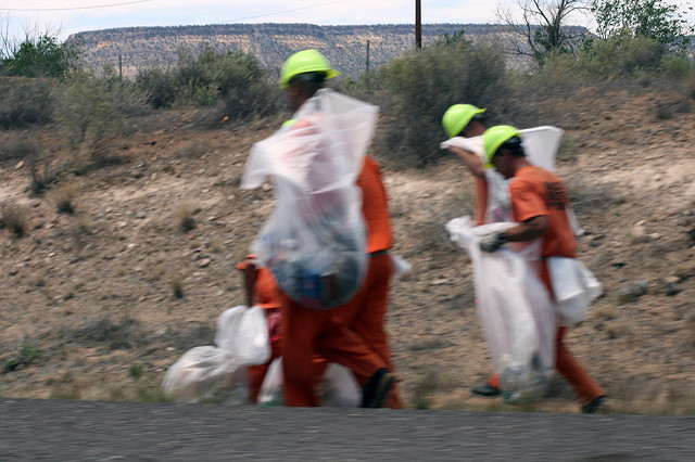 Incarcerated people in orange uniforms pick up trash by the side of a road.